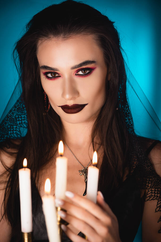 enfoque selectivo de la mujer joven con maquillaje negro y velo cerca de encender velas en azul - Foto, Imagen