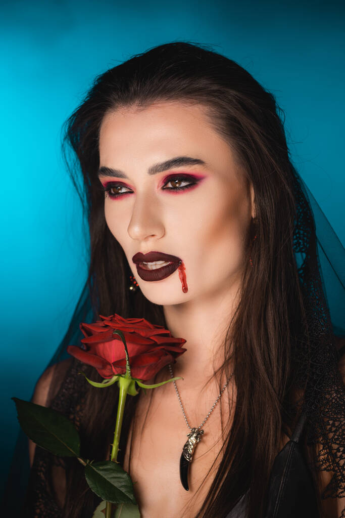ανατριχιαστική γυναίκα με αίμα στο πρόσωπο κοντά στο κόκκινο τριαντάφυλλο στο μπλε - Φωτογραφία, εικόνα