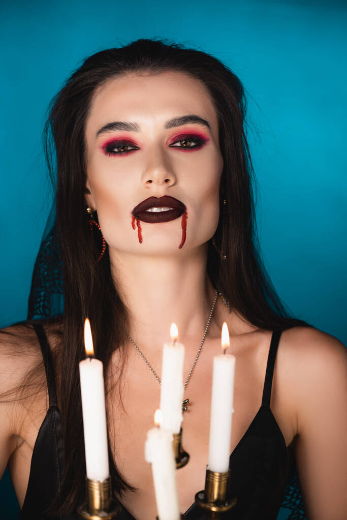 επιλεκτική εστίαση της γυναίκας με κόκκινο αίμα στο πρόσωπο κοντά στην καύση κεριών σε μπλε - Φωτογραφία, εικόνα