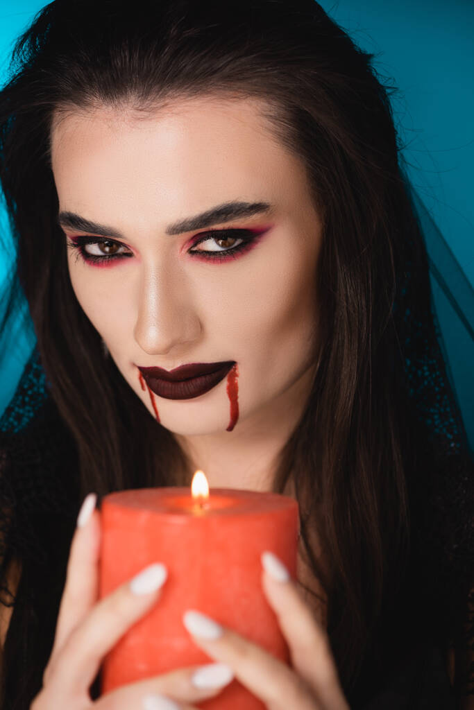 enfoque selectivo de la mujer morena con sangre en la cara sosteniendo vela ardiente en azul - Foto, Imagen
