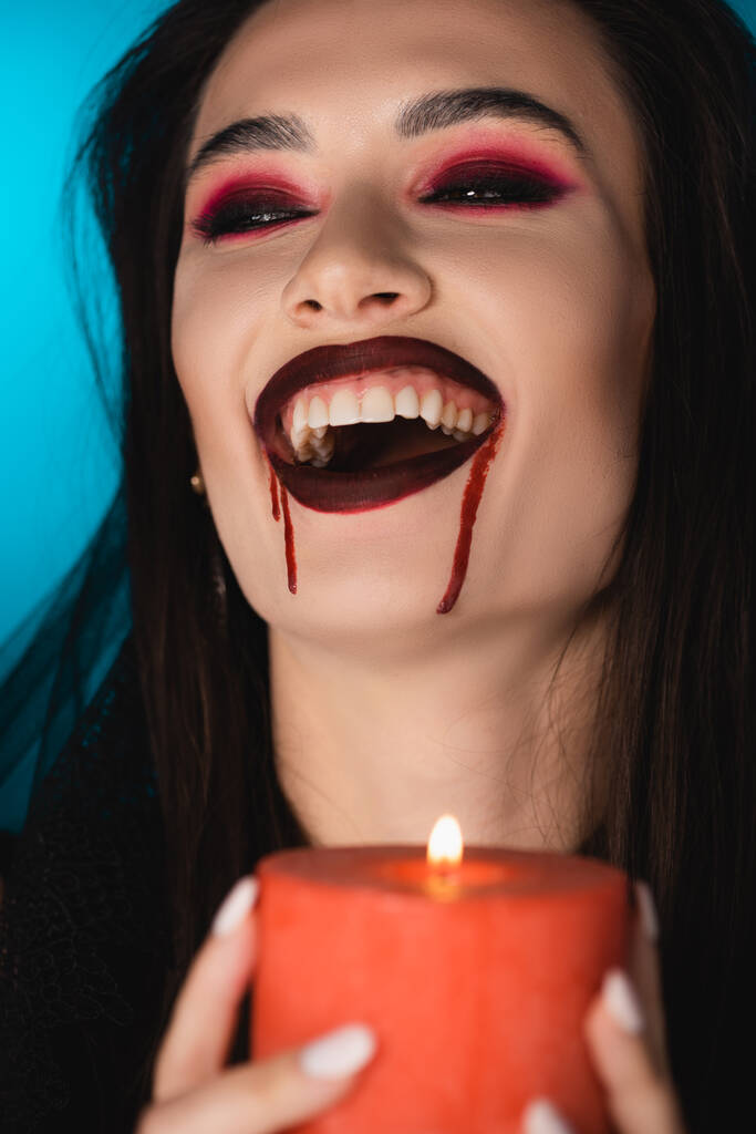 selectieve focus van brunette vrouw met bloed op gezicht met brandende kaars en lachen op blauw - Foto, afbeelding