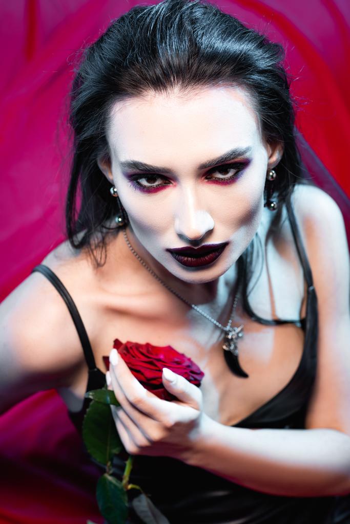 vue grand angle de la femme pâle avec du sang sur le visage regardant la caméra et tenant rose sur rouge - Photo, image