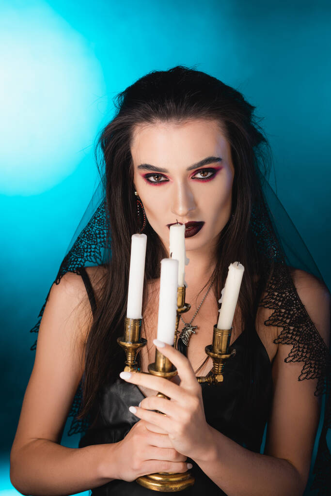 bleke vrouw met zwarte make-up met brandende kaarsen op blauw - Foto, afbeelding