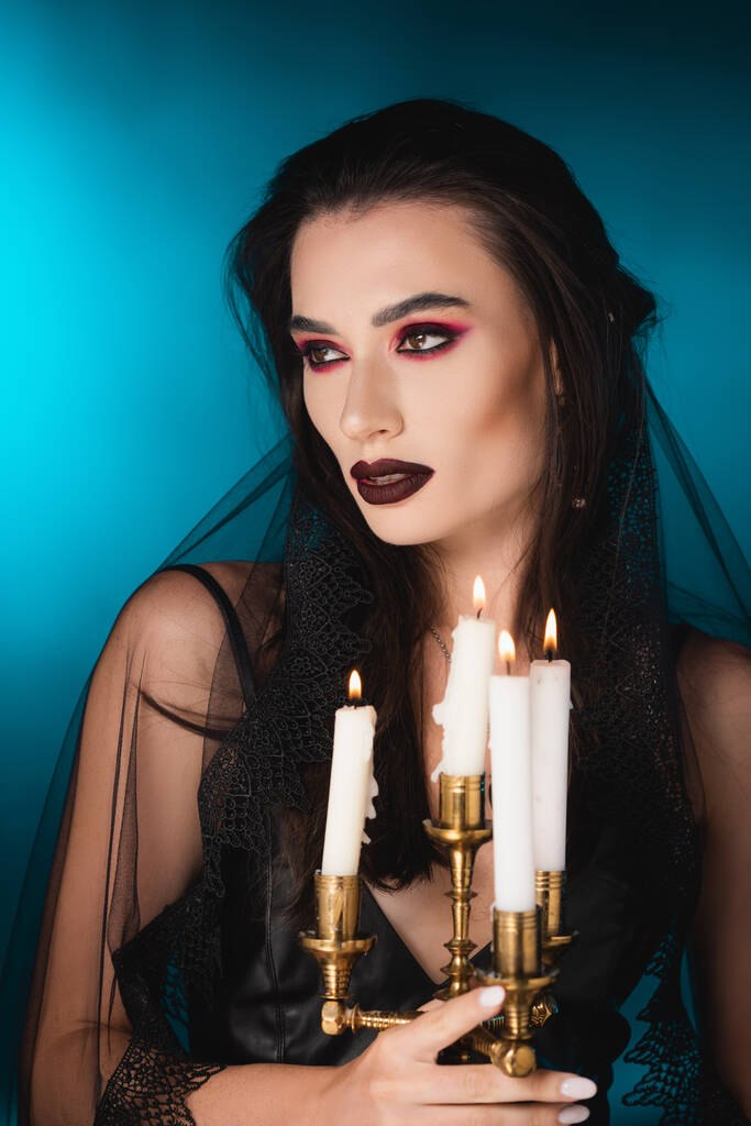 blasse Frau mit schwarzem Make-up, die wegschaut und brennende Kerzen auf blauem Grund hält - Foto, Bild