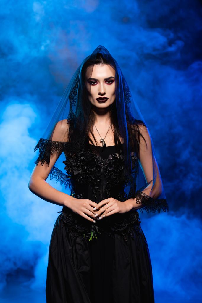 Braut in schwarzem Kleid und Schleier auf blau mit Rauch, Halloween-Konzept - Foto, Bild