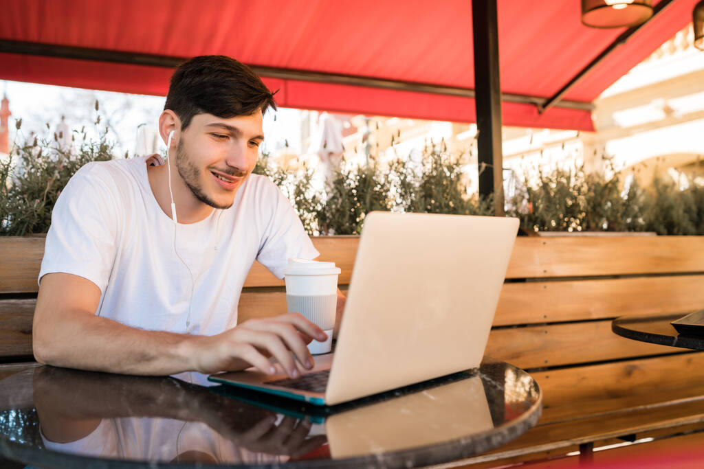 Πορτρέτο του νεαρού που χρησιμοποιεί το laptop του ενώ κάθεται σε καφετέρια. Έννοια τεχνολογίας και τρόπου ζωής. - Φωτογραφία, εικόνα