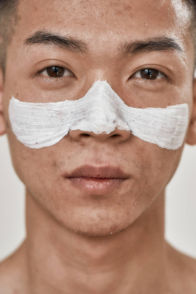 Detailní portrét mladého asijského muže s problematickou pletí a hyperpigmentací nanesenou maskou na jeho tváři, při pohledu na kameru izolovanou přes bílé pozadí. Krása, péče o pleť - Fotografie, Obrázek