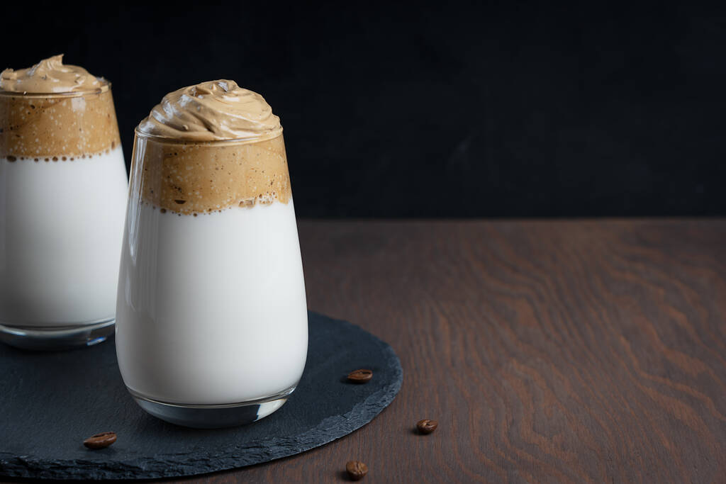 Glass of homemade dalgona drink made by whipping instant coffee powder, sugar and hot water with addition of cold milk on stony tray on dark wooden background at kitchen. Image with copy space - Photo, Image