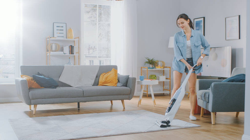 Young Beautiful Woman in Jeans Shirt and Shorts is Vacuum Cleaning a Carpet in a Bright Cozy Room at Home. She Uses a Modern Cordless Vacuum. Shes Happy and Cheerful. - Photo, Image