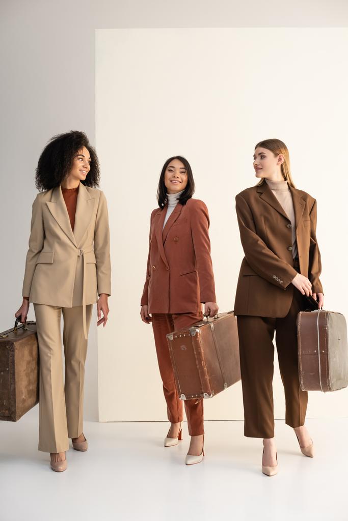 longitud completa de las mujeres interracial en trajes de moda con maletas en blanco - Foto, Imagen
