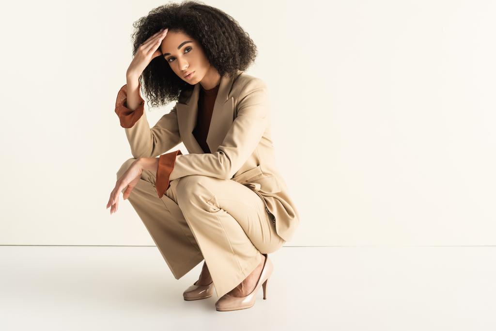 young and curly african american woman in stylish suit sitting on white - Photo, Image