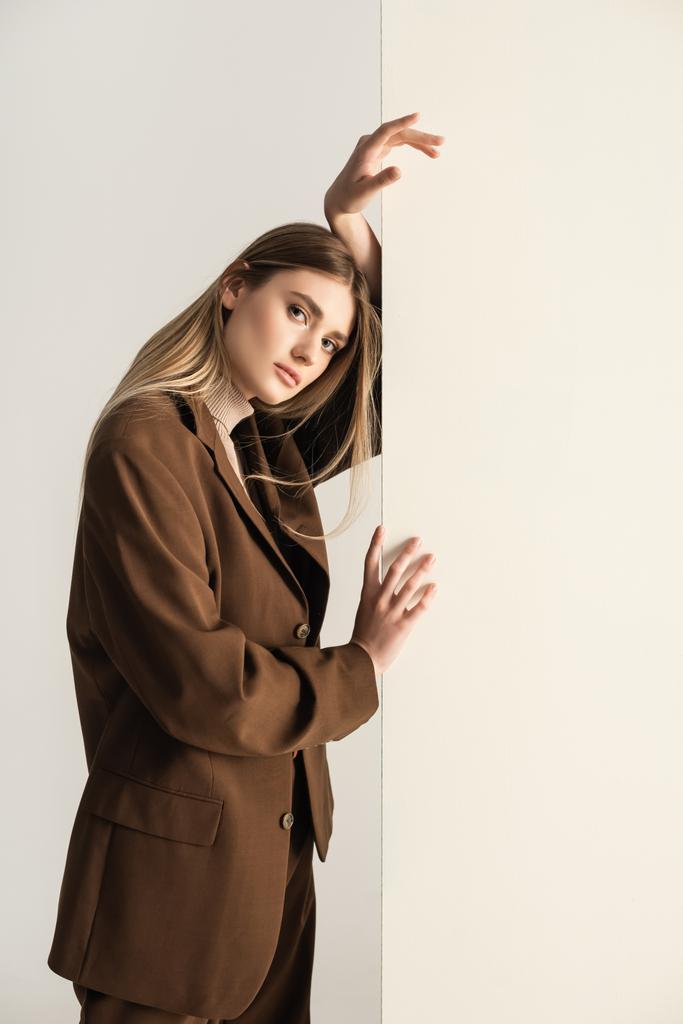 joven rubia en elegante traje marrón mirando a la cámara mientras se apoya en la pared en blanco - Foto, imagen