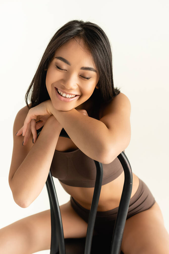 брюнетка азіатська жінка в бежевій білизні посміхається, позує на стільці ізольовано на білому
 - Фото, зображення