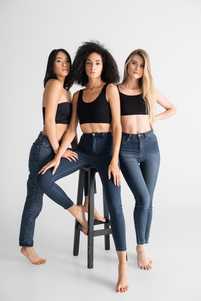 jóvenes interracial mujeres en denim jeans posando cerca de taburete en blanco - Foto, Imagen