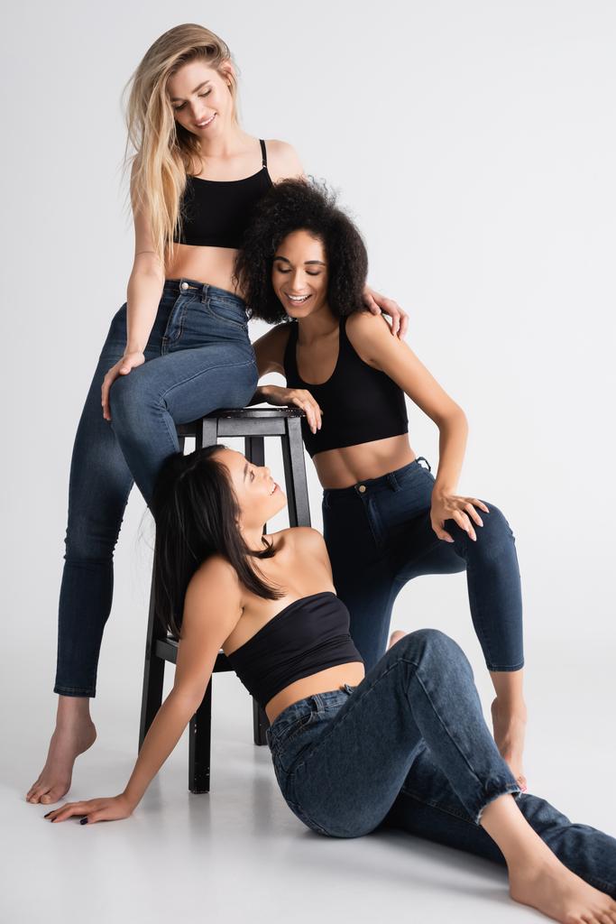 happy interracial women in denim jeans looking at each other while posing near stool on white - Photo, Image