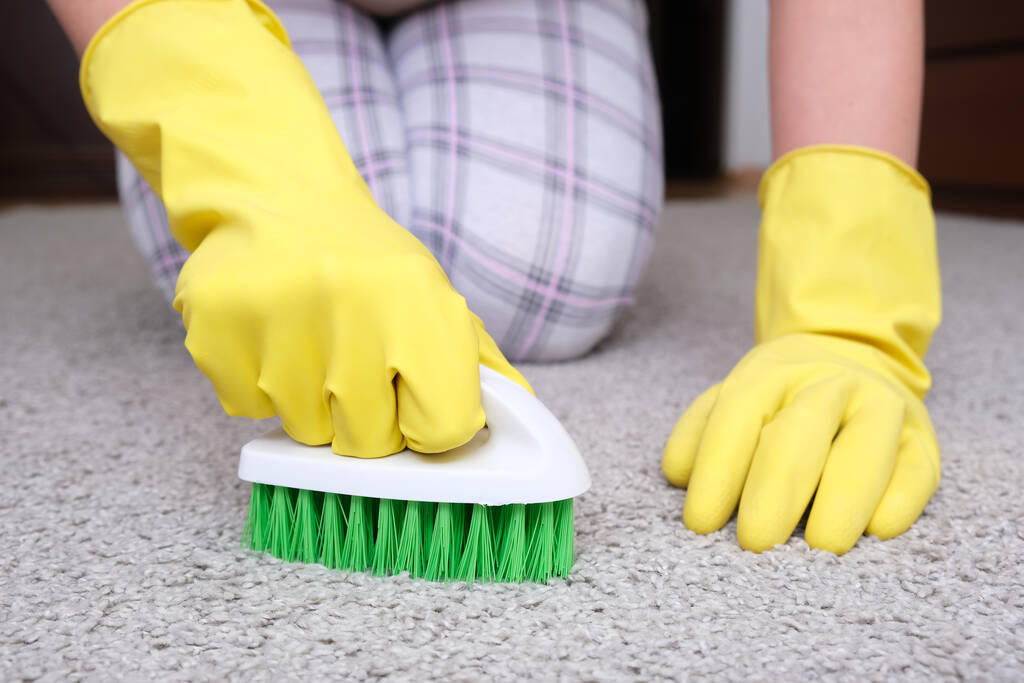 Frau in gelben Handschuhen mit grüner Bürste putzt und bürstet Teppich, entfernt Flecken und Wolle und erledigt Routineaufgaben - Foto, Bild