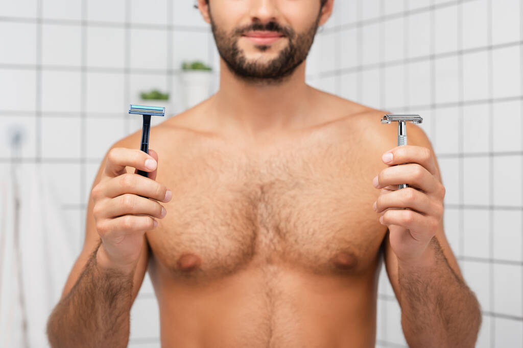 Oříznutý pohled na břitvy v rukou vousatého muže na rozmazaném pozadí v koupelně  - Fotografie, Obrázek