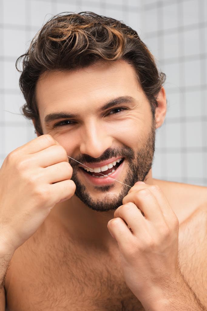 Sorridente uomo senza maglietta utilizzando filo interdentale mentre guardando la fotocamera in bagno  - Foto, immagini