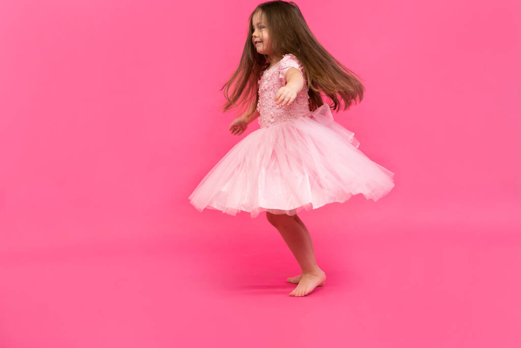 Το χαριτωμένο κοριτσάκι ονειρεύεται να γίνει μπαλαρίνα. Μικρό κορίτσι που χορεύει. Στούντιο σουτ πάνω από ροζ φόντο - Φωτογραφία, εικόνα