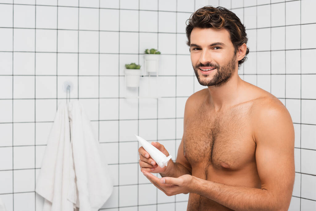 Homme torse nu souriant à la caméra tout en tenant tube avec crème cosmétique dans la salle de bain  - Photo, image