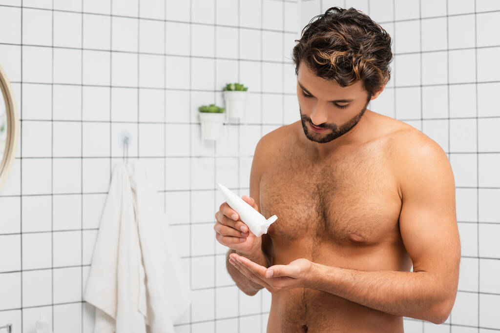 Muscular man squeezing cosmetic cream from tube in bathroom  - Photo, Image