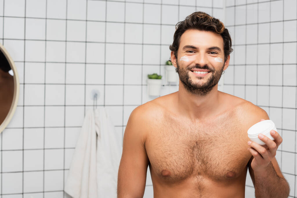 Homme torse nu souriant tenant pot avec crème cosmétique dans la salle de bain  - Photo, image