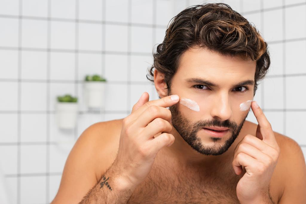 Бородатый мужчина наносит косметический крем на лицо в ванной комнате  - Фото, изображение