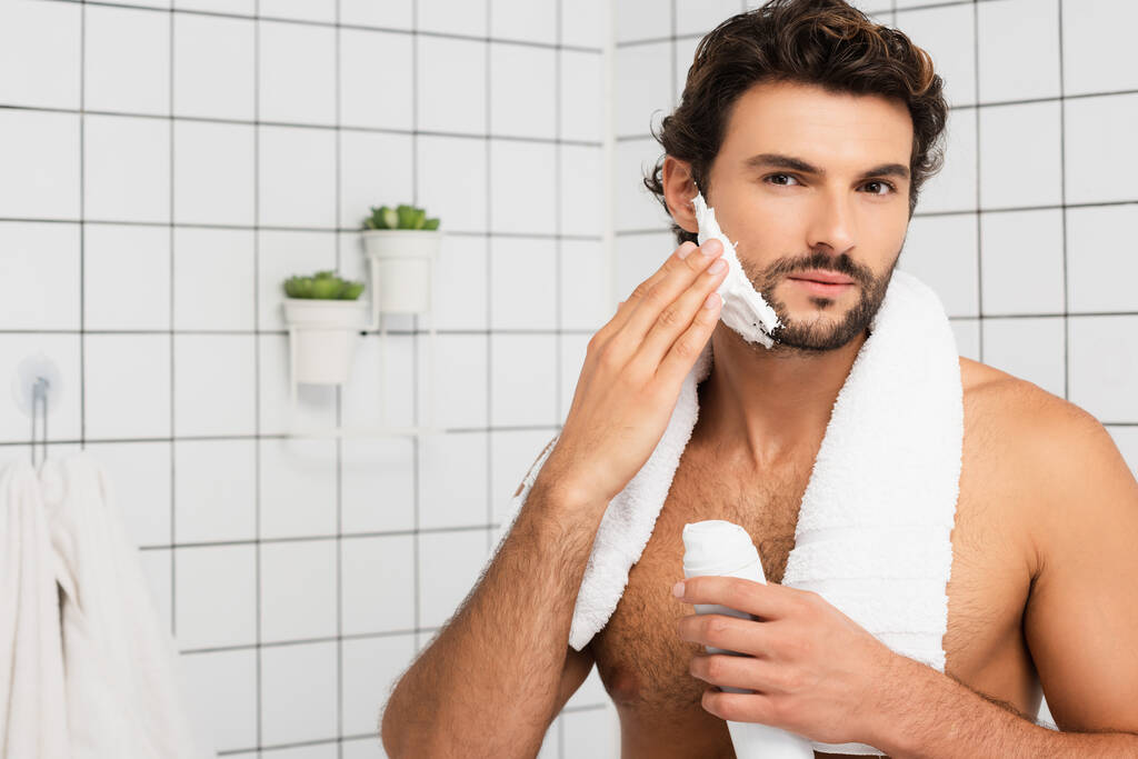 Hemdloser Mann schaut in Kamera, während er im Badezimmer Rasierschaum aufträgt  - Foto, Bild