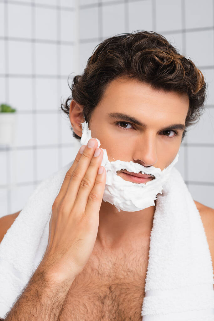 Morena hombre con toalla alrededor del cuello aplicando espuma de afeitar  - Foto, imagen