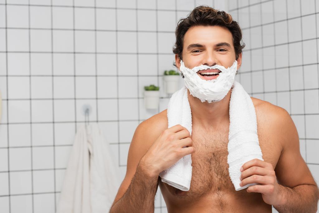 Wesoły mężczyzna bez koszuli z pianką do golenia na twarzy trzymający ręcznik w łazience  - Zdjęcie, obraz