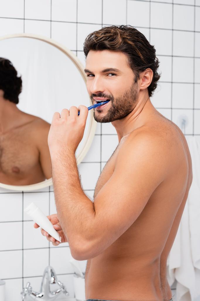 Безсоромний чоловік дивиться на камеру під час чищення зубів і тримає трубку зубною пастою у ванній
 - Фото, зображення