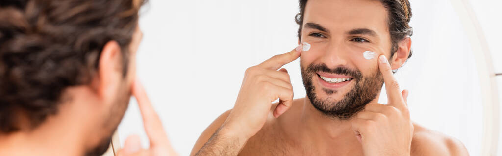 Homme souriant appliquant de la crème cosmétique sur le visage près du miroir sur le premier plan flou, bannière  - Photo, image