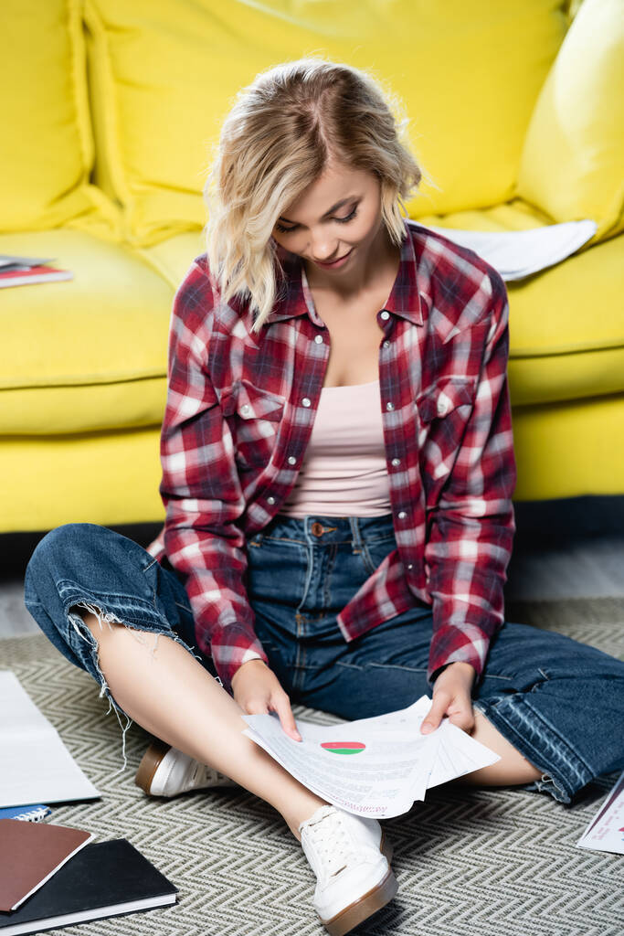 joven mujer rubia sentada en el suelo y mirando a través de documentos - Foto, imagen