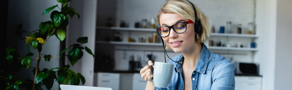 young blonde woman in eyeglasses and headphones drinking tea, horizontal banner - Foto, Imagen