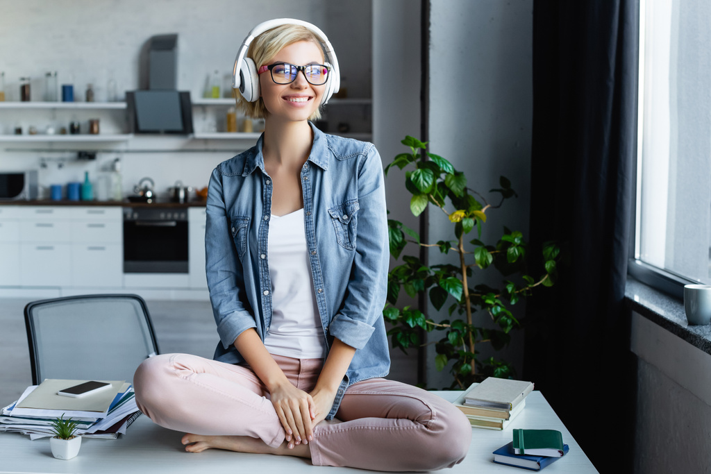 молодая блондинка в очках слушает музыку и сидит на столе - Фото, изображение