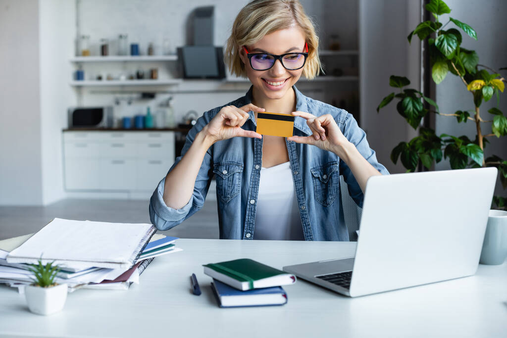 молодая блондинка в очках держит кредитку и делает покупки в интернете - Фото, изображение