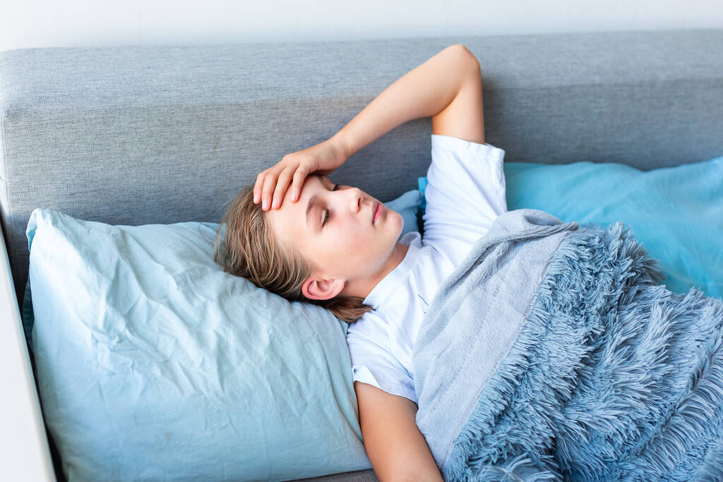 Sairas teini-ikäinen tyttö, jolla on korkea kuume ja päänsärky, makaa sängyssä tarkistamassa otsaansa. Pysy kotona koronavirusepidemian aikana, jos tunnet olosi sairaaksi - Valokuva, kuva