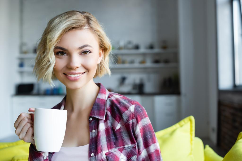 mujer rubia sonriente con camisa a cuadros sosteniendo taza blanca  - Foto, imagen