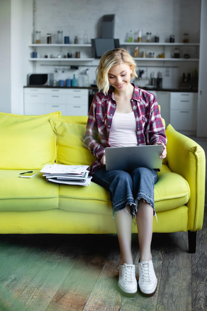 glimlachende blonde vrouw in geruite shirt werken vanuit huis met laptop op knieën - Foto, afbeelding