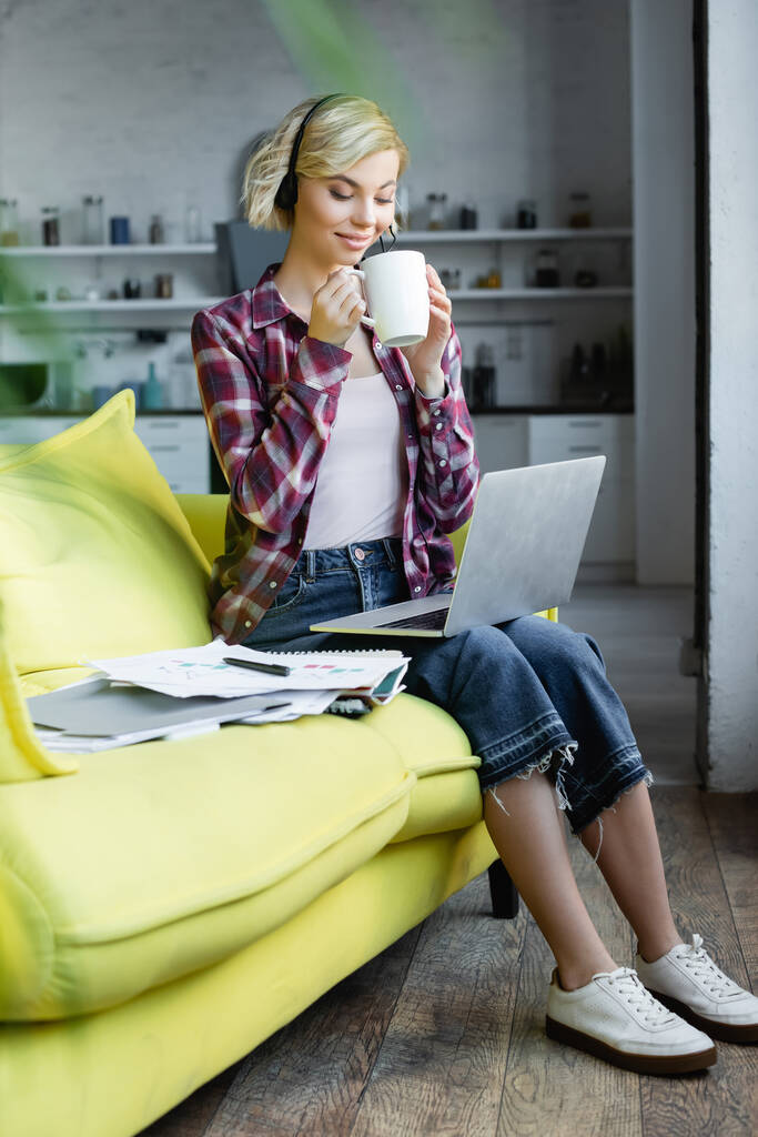 家で働いてお茶を飲むヘッドフォンの若いブロンドの女性 - 写真・画像
