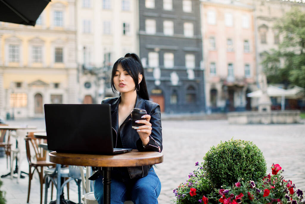 Horizontale outdoor shot van jonge aantrekkelijke Aziatische zakenvrouw, zitten aan tafel in cafe, met behulp van laptop op lunchpauze, het drinken van koffie. Vrouw freelancer werkt in het stadscafé - Foto, afbeelding