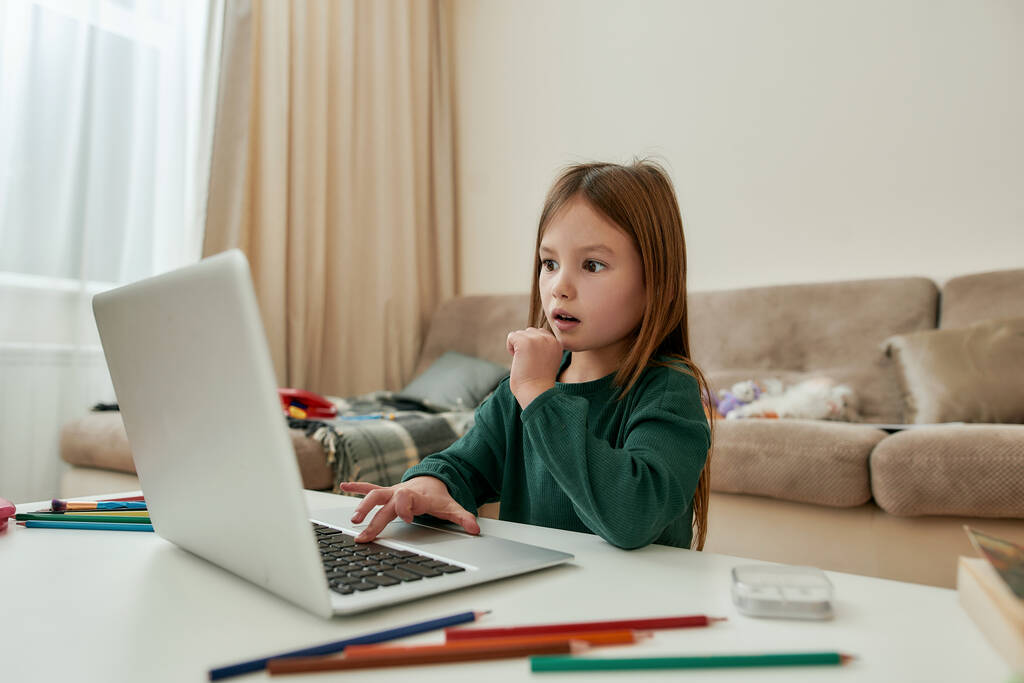 Egy aranyos kislány, aki csodálkozását fejezi ki azzal, hogy egy laptopot néz, amint egyedül ül az asztalnál a távoktatás alatt. - Fotó, kép