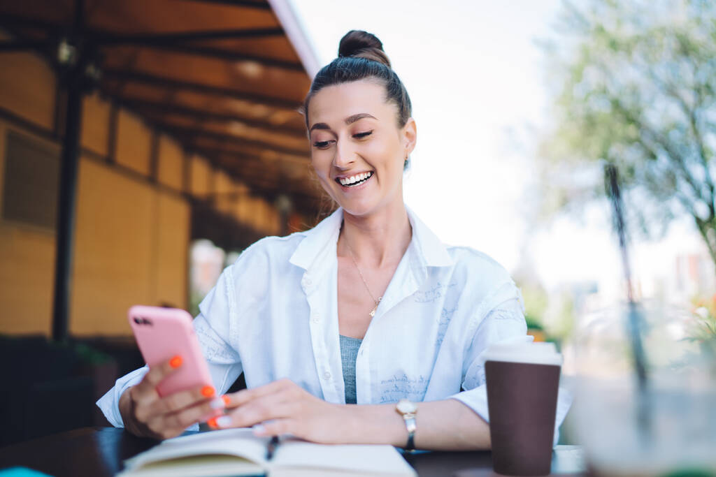 Mujer alegre en ropa casual sonriendo y mensajería en el teléfono inteligente mientras descansa en la cafetería al aire libre y beber café caliente para llevar - Foto, imagen