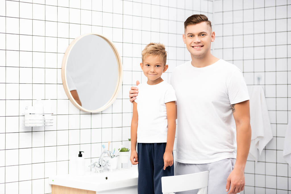 Улыбающийся отец обнимает сына, стоящего на стуле возле зеркала и раковины в ванной - Фото, изображение