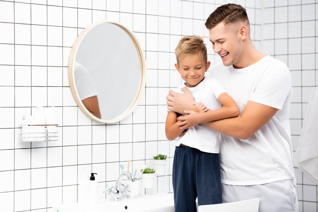 Mutlu baba oğluna sarılmış lavabonun yanında duran ve banyoda yuvarlak ayna olan. - Fotoğraf, Görsel