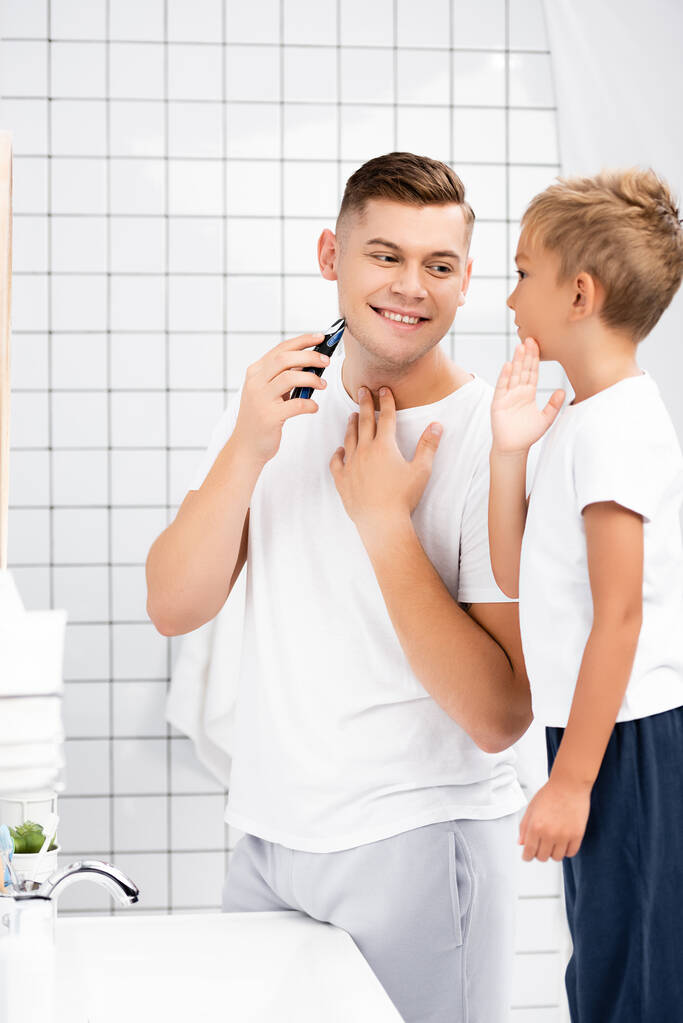Padre sorridente con rasoio elettrico in piedi vicino al figlio che tocca la guancia in bagno - Foto, immagini