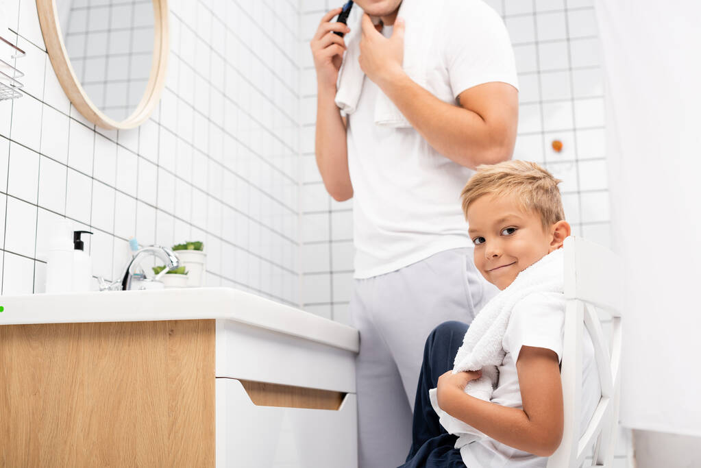 Pozitivní chlapec dívá na kameru, zatímco sedí na židli poblíž muže holení s elektrickou holicí strojek v koupelně - Fotografie, Obrázek