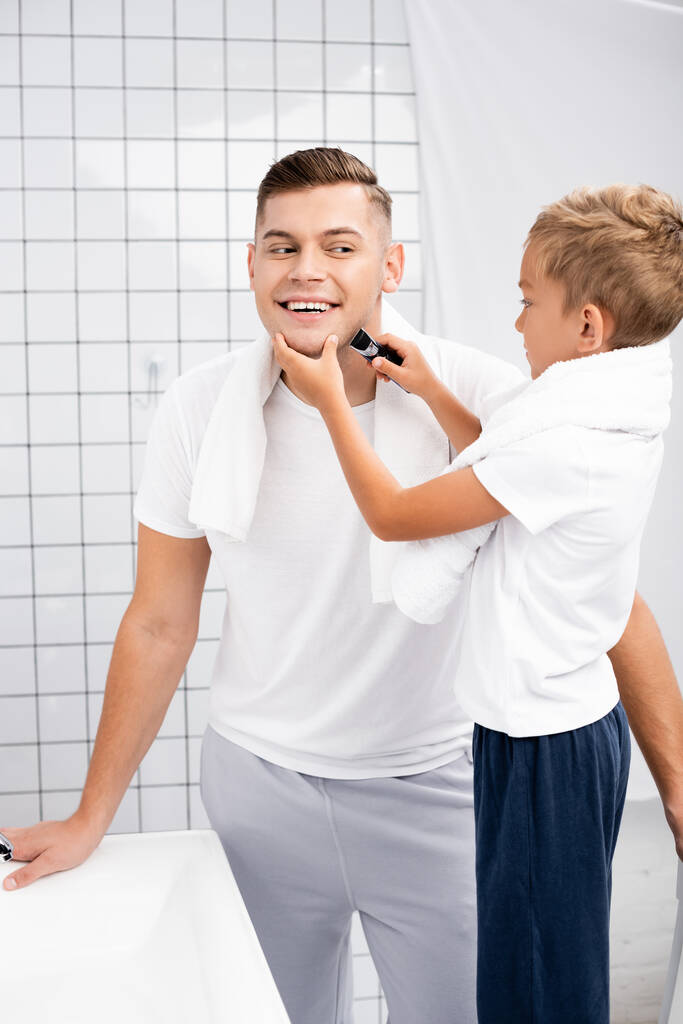 Сосредоточенный мальчик бреет щеку счастливого человека с электрической бритвой в ванной комнате - Фото, изображение