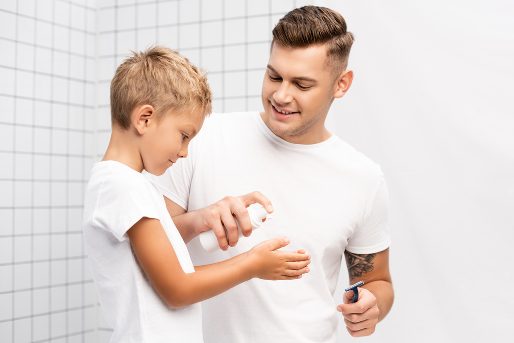 Sonriente padre con afeitadora de seguridad apretando espuma de afeitar en las manos del hijo en el baño - Foto, Imagen