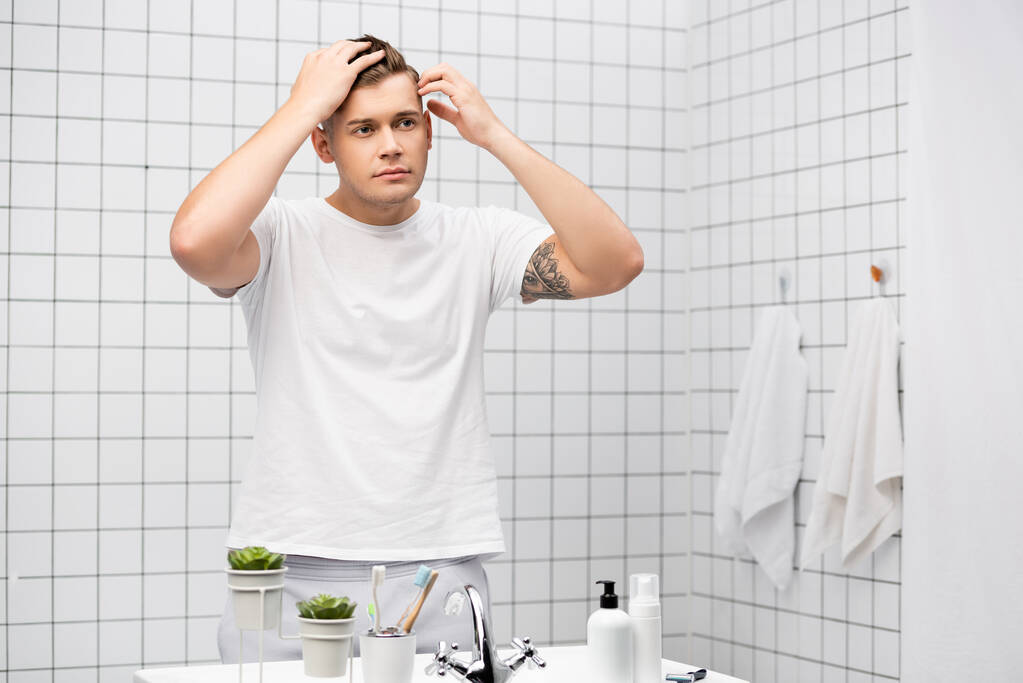 Genç yetişkin bir adam banyoda lavabonun yanında durup saçlarını kontrol ediyor. - Fotoğraf, Görsel